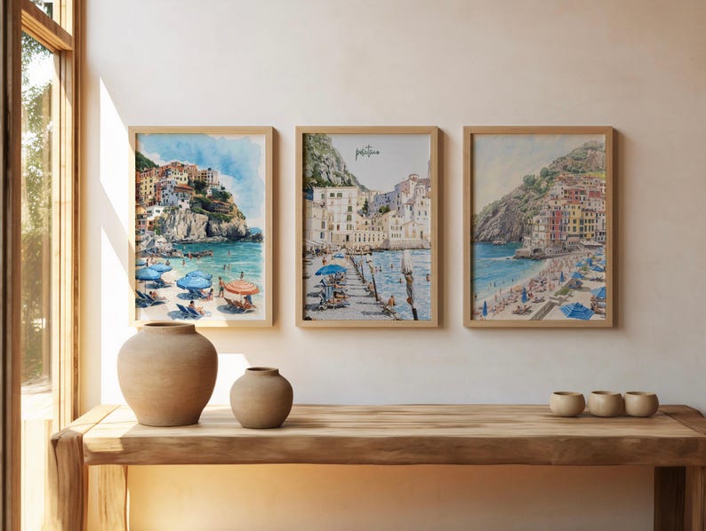 May include: Three framed watercolor prints of coastal scenes with buildings and beaches. The prints are in light wood frames. Two beige ceramic vases and three small beige ceramic bowls sit on a wooden table. The window frame is visible on the left.