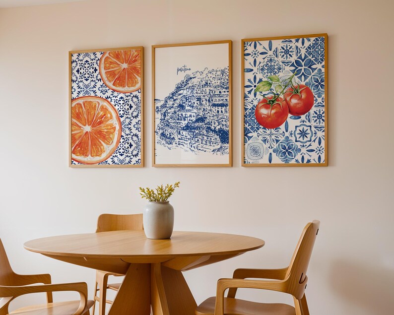 May include: Three framed art prints hang on a wall. The prints feature images of orange slices, a coastal town, and tomatoes. The prints have wooden frames and are displayed above a wooden table and chairs.