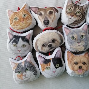 Puede incluir: Una colecci&oacute;n de nueve almohadas de peluche de mascotas con diferentes razas de perros y gatos. Las almohadas son blancas con im&aacute;genes coloridas de las caras de los animales. Las almohadas est&aacute;n dispuestas en semic&iacute;rculo sobre una superficie blanca.