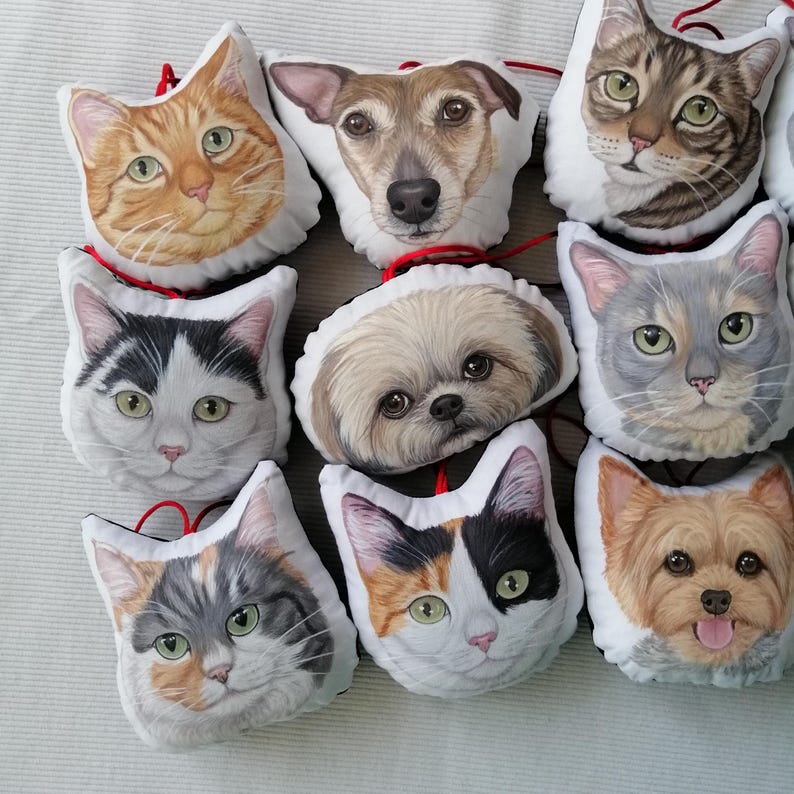 Puede incluir: Una colecci&oacute;n de nueve almohadas de peluche de mascotas con diferentes razas de perros y gatos. Las almohadas son blancas con im&aacute;genes coloridas de las caras de los animales. Las almohadas est&aacute;n dispuestas en semic&iacute;rculo sobre una superficie blanca.