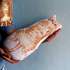 K&ouml;nnte beinhalten: Ein wei&szlig;es Kissen mit einem handgemalten Design eines orangefarbenen Tabby-Katers. Die Katze liegt auf dem R&uuml;cken mit den Pfoten in der Luft.
