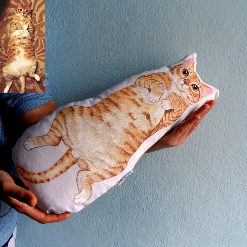 K&ouml;nnte beinhalten: Ein wei&szlig;es Kissen mit einem handgemalten Design eines orangefarbenen Tabby-Katers. Die Katze liegt auf dem R&uuml;cken mit den Pfoten in der Luft.