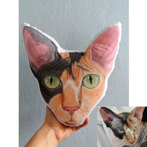 K&ouml;nnte beinhalten: Ein Pl&uuml;schkissen in Form eines Katzenkopfes. Die Katze hat schwarzes, orangefarbenes und wei&szlig;es Fell und gr&uuml;ne Augen. Das Kissen ist aus weichem Stoff und perfekt zum Kuscheln.