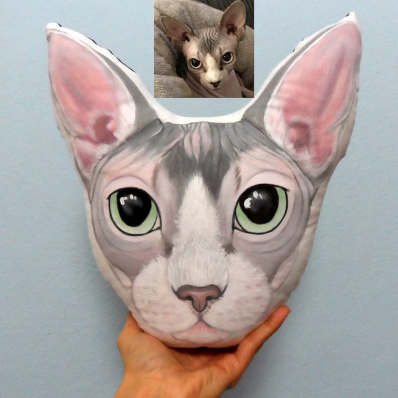 K&ouml;nnte beinhalten: Ein handbemaltes Kissen mit einem Sphynx-Kater mit gr&uuml;nen Augen und einem grau-wei&szlig;en Fell. Das Kissen ist aus weichem Stoff gefertigt und ein einzigartiges und pers&ouml;nliches Geschenk.