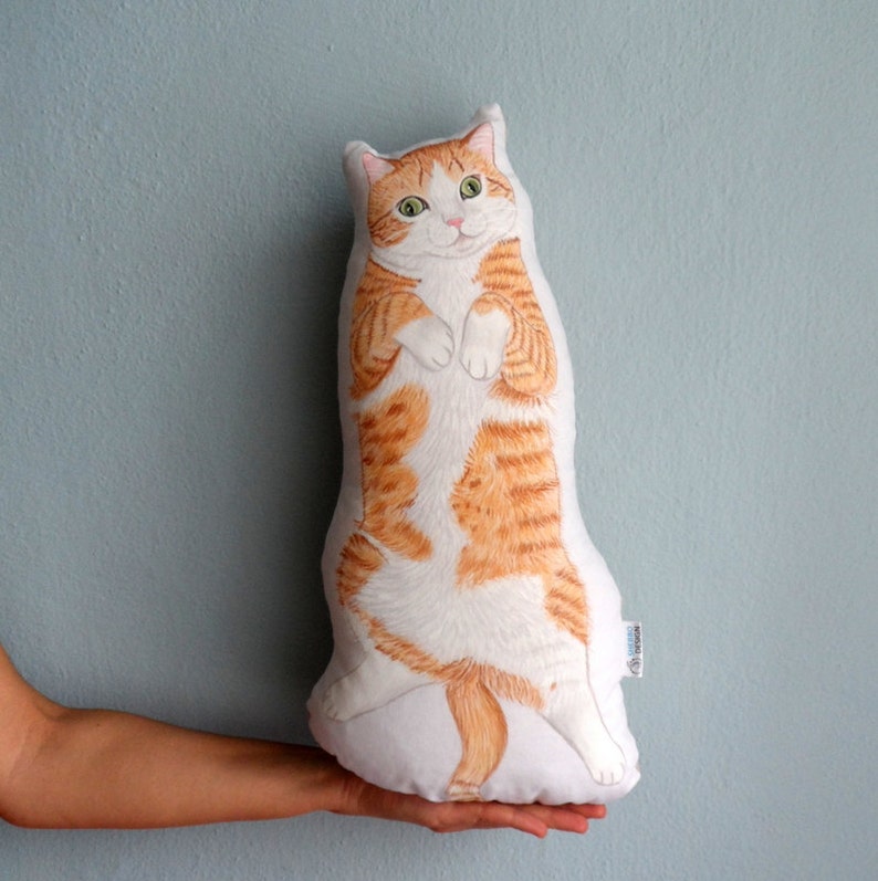 K&ouml;nnte beinhalten: Ein wei&szlig;es Kissen mit einem handgemalten orange-wei&szlig;en Kater, der auf seinen Hinterbeinen steht. Die Katze hat gr&uuml;ne Augen und eine rosa Nase.