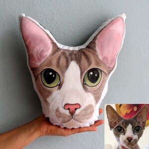 K&ouml;nnte beinhalten: Ein individuelles Kissen mit einem handgemalten Portr&auml;t einer braunen und wei&szlig;en haarlosen Katze mit gr&uuml;nen Augen. Das Kissen ist wei&szlig; mit einem schwarzen Umriss und wird in der Hand einer Person gehalten.