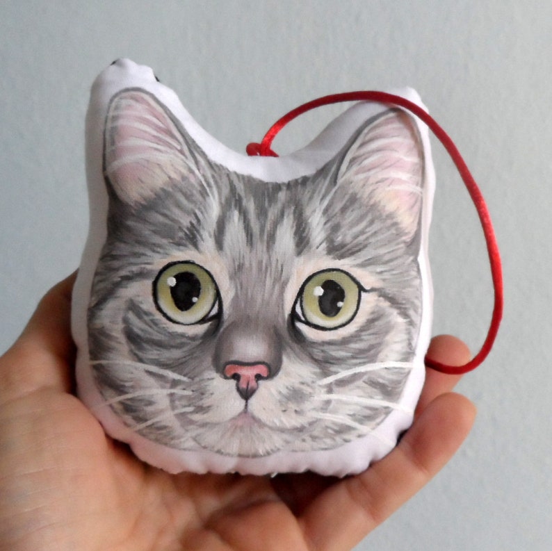 Puede incluir: Un adorno de cara de gato gris y blanco con una cinta roja. El gato tiene ojos verdes y una nariz rosa.
