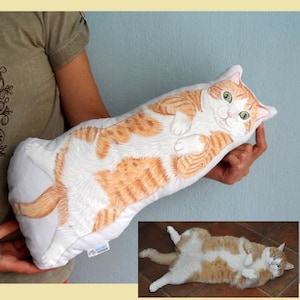 K&ouml;nnte beinhalten: Ein ma&szlig;geschneidertes Kissen mit einer handgemalten Illustration einer rot-wei&szlig;en Katze, die auf dem R&uuml;cken liegt. Das Kissen ist wei&szlig; mit einem blauen Etikett, auf dem "Custom Design" steht.