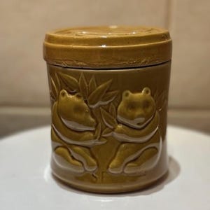 May include: A ceramic jar with a lid, in a glossy, golden-brown hue. The jar is decorated with embossed frog and bamboo leaf designs. The cylindrical jar is on a white surface. The jar is approximately 10 cm tall.