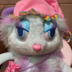 Puede incluir: Un peluche con cuerpo azul claro, detalles morados y ojos azules grandes. Lleva un gorro rosa con ribete arcoíris y tiene una nariz rosa. Una bolsa rosa con diseño de arcoíris cuelga de su cuello. El juguete tiene largas pestañas oscuras.