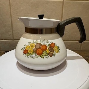 May include: A vintage white and tan coffee pot with a black handle and lid. The pot features a floral design with oranges, yellow flowers, and green leaves. A gold band encircles the middle of the pot.