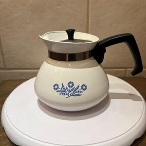 May include: A white teapot with a rounded body and a black handle. The teapot features a blue floral pattern on the front, a metallic band around the neck, and a black lid knob. The design is simple, with a wide base and a curved spout for pouring.