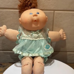 以下が含まれることがあります： 丸い顔、青い目、赤褐色の髪の毛をした、ヴィンテージのキャベツ人形。人形は、レースのトリムとマッチするおむつが付いた水色のドレスを着ています。人形の腕と脚は柔らかい生地で作られています。