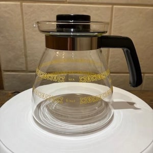 Puede incluir: Una taza de cerámica blanca con un asa negra y el texto "But First, Coffee" en negrita negra. La taza se encuentra sobre un fondo borroso, sugiriendo un ambiente acogedor.