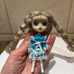 May include: A small doll with blonde, curly pigtails and large blue eyes. The doll wears a turquoise dress with white lace trim and a white ribbon. The doll is held in a hand.