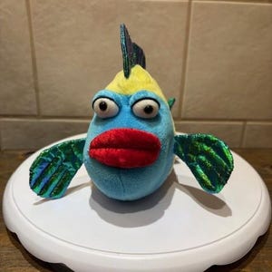 May include: A plush toy fish with a cartoon design. The fish has a blue body, yellow head, large googly eyes, and a red plush mouth. Its fins are green with an iridescent finish. The toy is on a white, round surface.