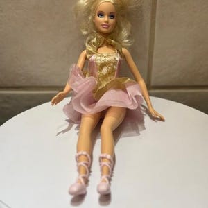 Può includere: Bambola bionda in un completo da ballerina rosa e oro. La bambola ha gli occhi azzurri e indossa scarpette da ballo rosa. L'abito ha un corpetto dorato e una gonna rosa a strati. La bambola è posata su una superficie bianca.