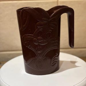 Peut inclure: Une tasse en plastique marron foncé avec une poignée et un motif de visage de lapin. La tasse a une large ouverture et une poignée incurvée. Le visage du lapin est en relief sur le devant.
