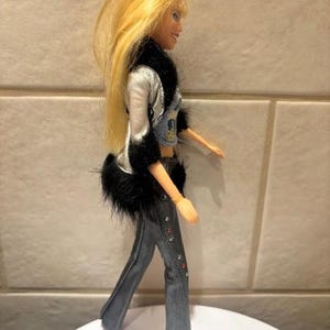Peut inclure: Une poup&eacute;e aux longs cheveux blonds, v&ecirc;tue d'une veste argent&eacute;e avec une bordure en fausse fourrure noire, d'un crop top en jean et d'un pantalon &eacute;vas&eacute; en jean. Le pantalon est orn&eacute; de strass rouges et transparents. La poup&eacute;e est sur une surface blanche.