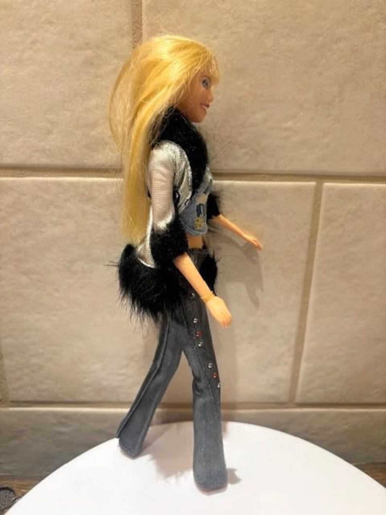 Peut inclure: Une poup&eacute;e aux longs cheveux blonds, v&ecirc;tue d'une veste argent&eacute;e avec une bordure en fausse fourrure noire, d'un crop top en jean et d'un pantalon &eacute;vas&eacute; en jean. Le pantalon est orn&eacute; de strass rouges et transparents. La poup&eacute;e est sur une surface blanche.
