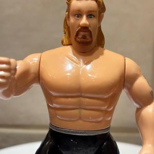May include: A vintage action figure of a muscular man with tan skin, blonde hair, and a goatee. The figure is wearing black trousers and has a silver belt. The figure is posed with one arm extended.