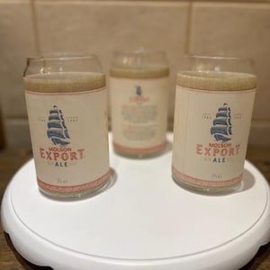 May include: Three clear glass beer can-shaped glasses with the Molson Export Ale logo on a white label. The logo features a blue ship and red text. The glasses are filled with a light-colored liquid and sit on a white, round tray.