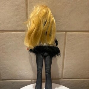Peut inclure: Une poup&eacute;e aux longs cheveux blonds, v&ecirc;tue d'une tenue noire. L'ensemble de la poup&eacute;e comprend un pantalon noir et un haut avec une bordure en fourrure noire. La poup&eacute;e est debout sur une surface blanche.