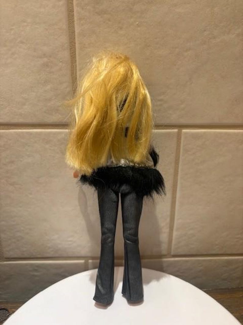 Peut inclure: Une poup&eacute;e aux longs cheveux blonds, v&ecirc;tue d'une tenue noire. L'ensemble de la poup&eacute;e comprend un pantalon noir et un haut avec une bordure en fourrure noire. La poup&eacute;e est debout sur une surface blanche.