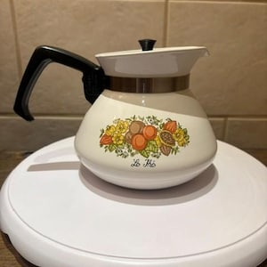 Puede incluir: Tetera vintage de color crema con asa y tapa negras. La tetera presenta una banda marrón y un diseño floral con el texto "Le Thé". La tetera está sobre una superficie blanca.