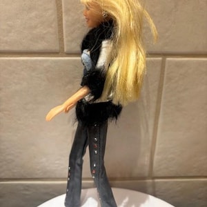 Peut inclure: Poup&eacute;e mannequin aux longs cheveux blonds, v&ecirc;tue d'une veste noire et blanche avec une bordure en fausse fourrure noire, d'un haut en jean et d'un pantalon gris fonc&eacute; avec des accents rouges et argent&eacute;s. La poup&eacute;e est sur une surface blanche.