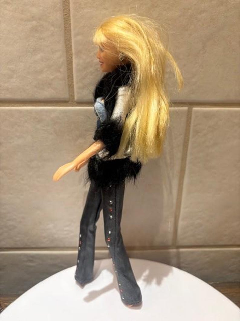 Peut inclure: Poup&eacute;e mannequin aux longs cheveux blonds, v&ecirc;tue d'une veste noire et blanche avec une bordure en fausse fourrure noire, d'un haut en jean et d'un pantalon gris fonc&eacute; avec des accents rouges et argent&eacute;s. La poup&eacute;e est sur une surface blanche.