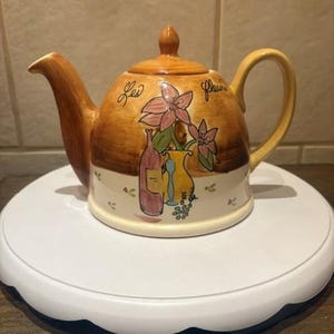 May include: A ceramic teapot with a tan and cream finish, featuring a hand-painted design of pink flowers, a wine bottle, and a glass. The text Les fleurs is written in black script on the upper portion. It has a curved handle and spout.