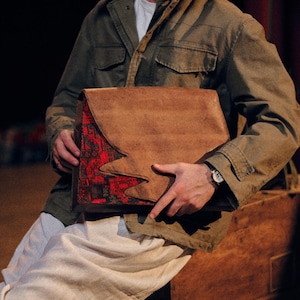 May include: A brown leather messenger bag with a large flap and a jagged edge design revealing a red and black patterned fabric underneath. The bag features a structured rectangular shape and is held by a person wearing a green jacket and white trousers.