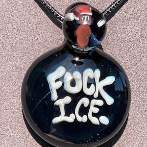 Può includere: Collana con pendente in vetro nero con le parole "FUCK ICE" in lettere bianche. Il pendente ha una forma rotonda con una piccola perla di vetro nella parte superiore con un personaggio dei cartoni animati con un cappello rosso.