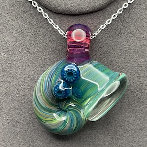 Op de afbeelding: Een handgemaakte glazen hangerketting. De hanger heeft een wervelend ontwerp in groen-, blauw- en geeltinten, met twee kleine blauwe bollen. De bovenkant is paars en rood. De ketting heeft een zilveren ketting.