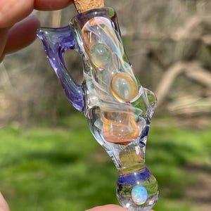 May include: A clear glass bottle with a cork stopper, featuring a swirling design of orange, green, and white. The bottle has a purple handle and a blue and white base with an opal-like stone. The bottle is held in a hand.