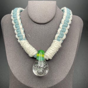 May include: A handmade necklace featuring a braided cord in white and light blue. The pendant is a clear glass teardrop shape with bubbles, topped with a green and yellow glass bead. The necklace is displayed on a gray bust.