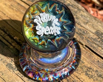 May include: A glass paperweight with a swirling blue, green, and yellow pattern. The top of the paperweight has a white inscription that reads "Nature Speaks".