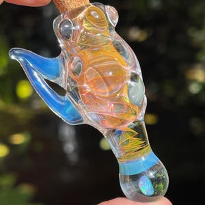 May include: A clear glass pendant shaped like a small bottle with a cork stopper. The glass features swirling orange and yellow internal patterns, a bright blue side accent, and a small iridescent opal inlay at the rounded base of the piece.