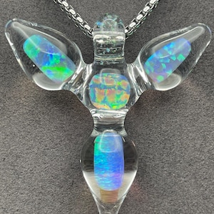 May include: A clear glass pendant shaped like a stylized figure with wings, featuring four embedded iridescent opal-like stones that display flashes of blue, green, and orange. The pendant hangs from a silver-toned metal box chain necklace.