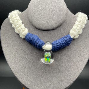 May include: A necklace featuring a central clear glass pendant with an internal iridescent green and blue pattern. The pendant is suspended from a cord wrapped in dark blue yarn, which transitions into thick, white, braided macrame-style cord.