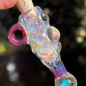 May include: A glass pendant featuring an iridescent, bubbled texture with swirling blue and purple hues. The piece includes a circular loop on the side, a cork stopper at the top, and a small, embedded blue opal-like stone at the rounded base.