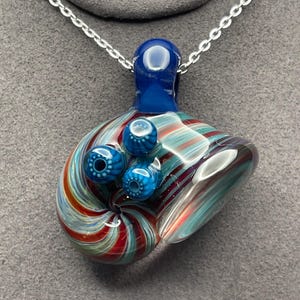 May include: A handcrafted glass pendant necklace with a swirling design of red, blue, and teal stripes. The pendant features three blue, spherical accents and a blue loop for the silver chain.