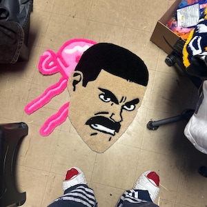 May include: A handmade rug featuring a stylized face with a black mustache and hair, and a pink shape. The rug is placed on a tiled floor. The image also shows a person's feet wearing white sneakers with red accents and blue and white striped pants.