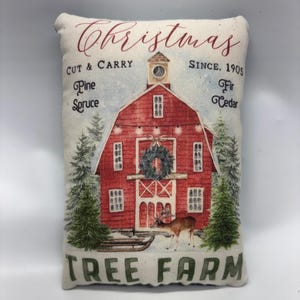 May include: A rectangular Christmas-themed pillow featuring a red barn illustration with a deer, Christmas tree, and the words "Christmas Tree Farm." The pillow also includes the text "Cut & Carry" and "Since 1905."