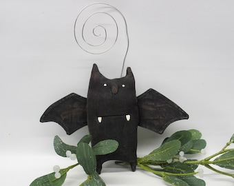 May include: A black felt bat with white teeth and eyes, hanging from a silver wire loop. The bat is sitting on a sprig of green mistletoe with white berries.