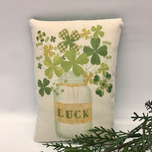 May include: A white pillow with a green shamrock design. The shamrocks are in a glass jar with the word "LUCK" written on it.