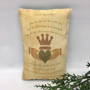 May include: A beige pillow with green trim features an Irish blessing printed in brown text. The text reads "May the road rise up to meet you, May the wind always be at your back, May the sun shine warm upon your face, and rains fall soft upon your fields, And until we meet again, May God hold you in the Palm of His hand." The pillow also features a green shamrock with a gold crown above it.