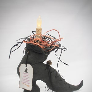 A handmade black witch's boot with a pointed toe and a black lace-up closure. The boot has a small bell on the side and a candle in a nest of raffia on top. A white tag with pink text hangs from the boot. The text on the tag reads: 'When witches go riding, and black cats are seen, the moon laughs and whispers, 'tis near Halloween.'