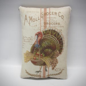 May include: A rectangular off-white throw pillow featuring a vintage-style graphic of a turkey carrying a basket of wheat and fruit. The text reads A MOLL GROCER CO. ESTABLISHED 1858 IMPORTERS AND WHOLESALE GROCERS, WINES, LIQUORS & CIGARS.